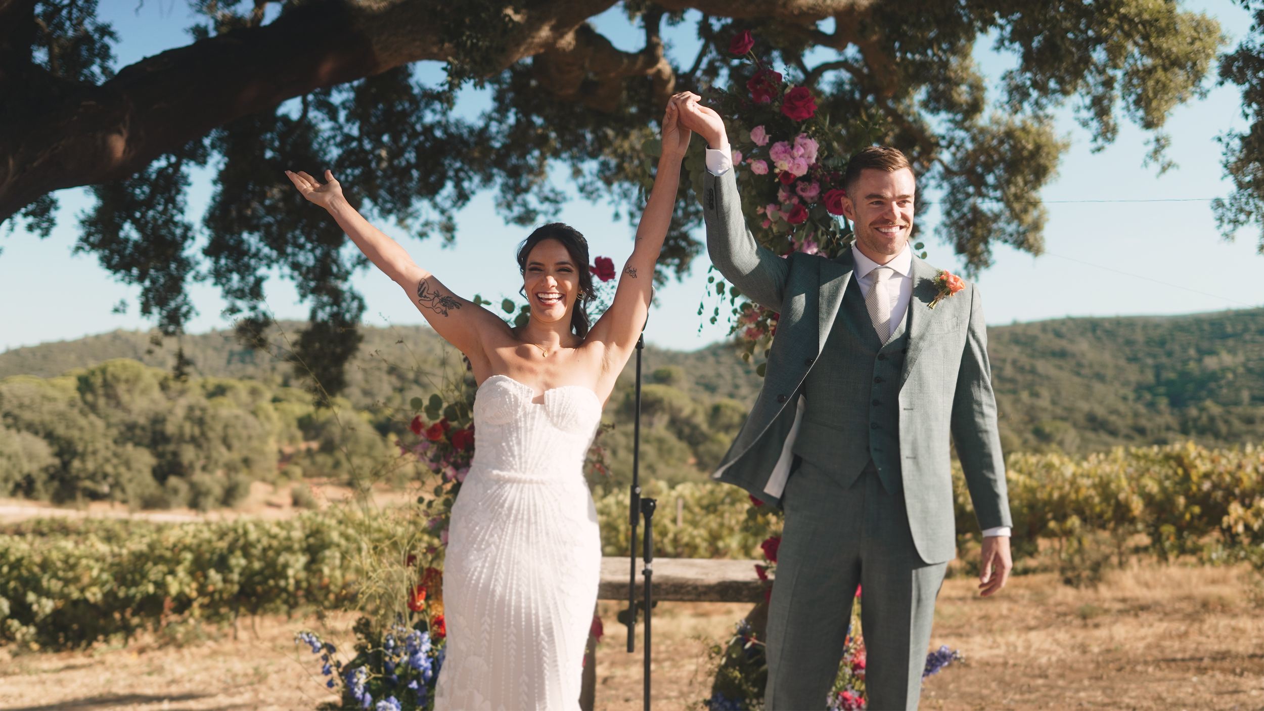 A smiling bride and groom stand outdoors beneath a large tree, holding hands raised in celebration after their wedding ceremony. The bride wears a white strapless wedding gown, and the groom is dressed in a gray suit with a boutonniere. Flowers and greenery decorate the background.