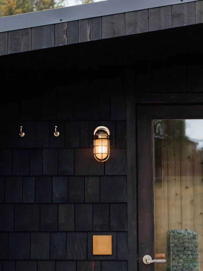 Close-up of a black wooden exterior wall with an outdoor wall light fixture and two hooks, with a door and glass window reflecting the outdoors.