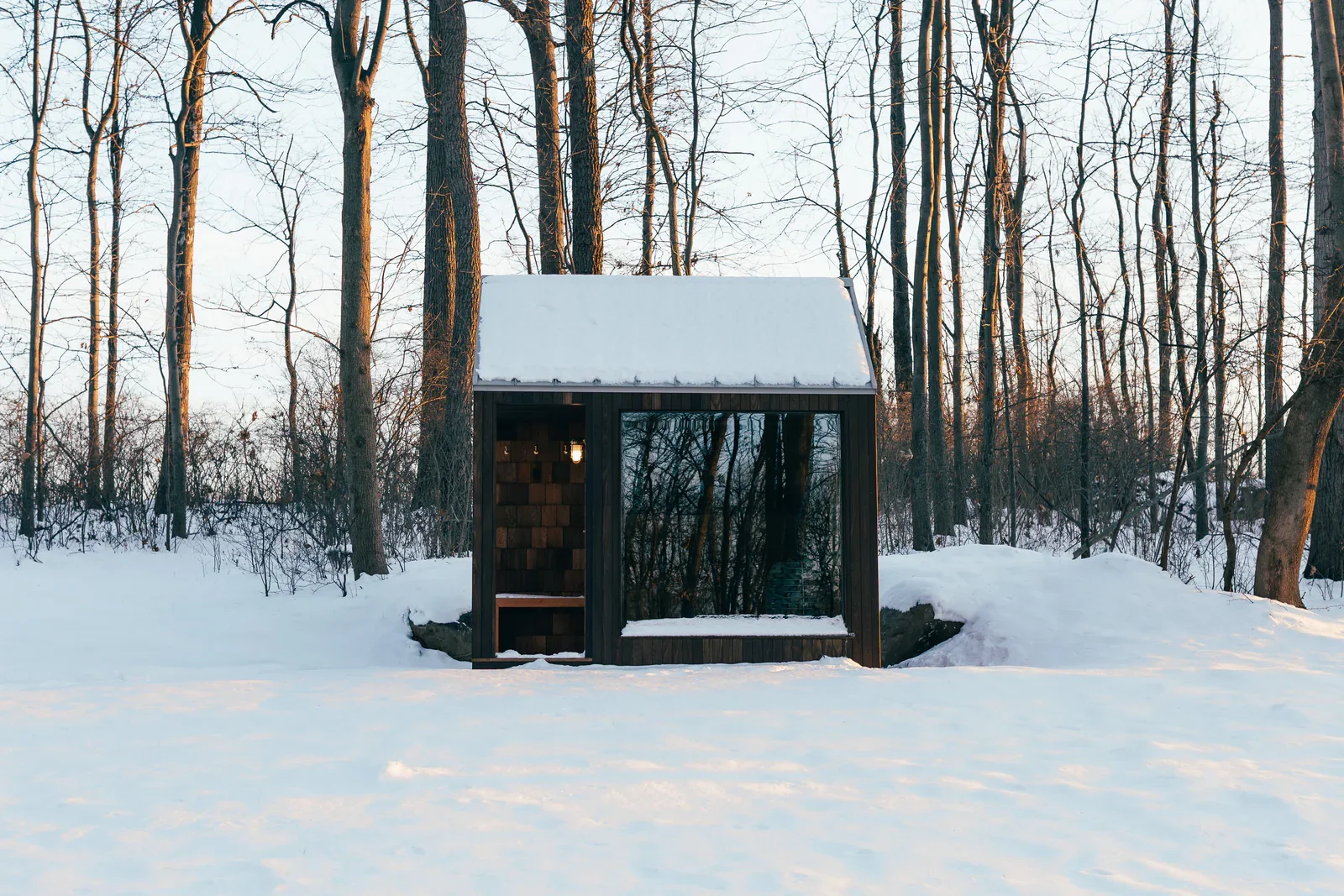 A small modern shelter or tiny house with a snow-covered roof located in a snowy forest with leafless trees.