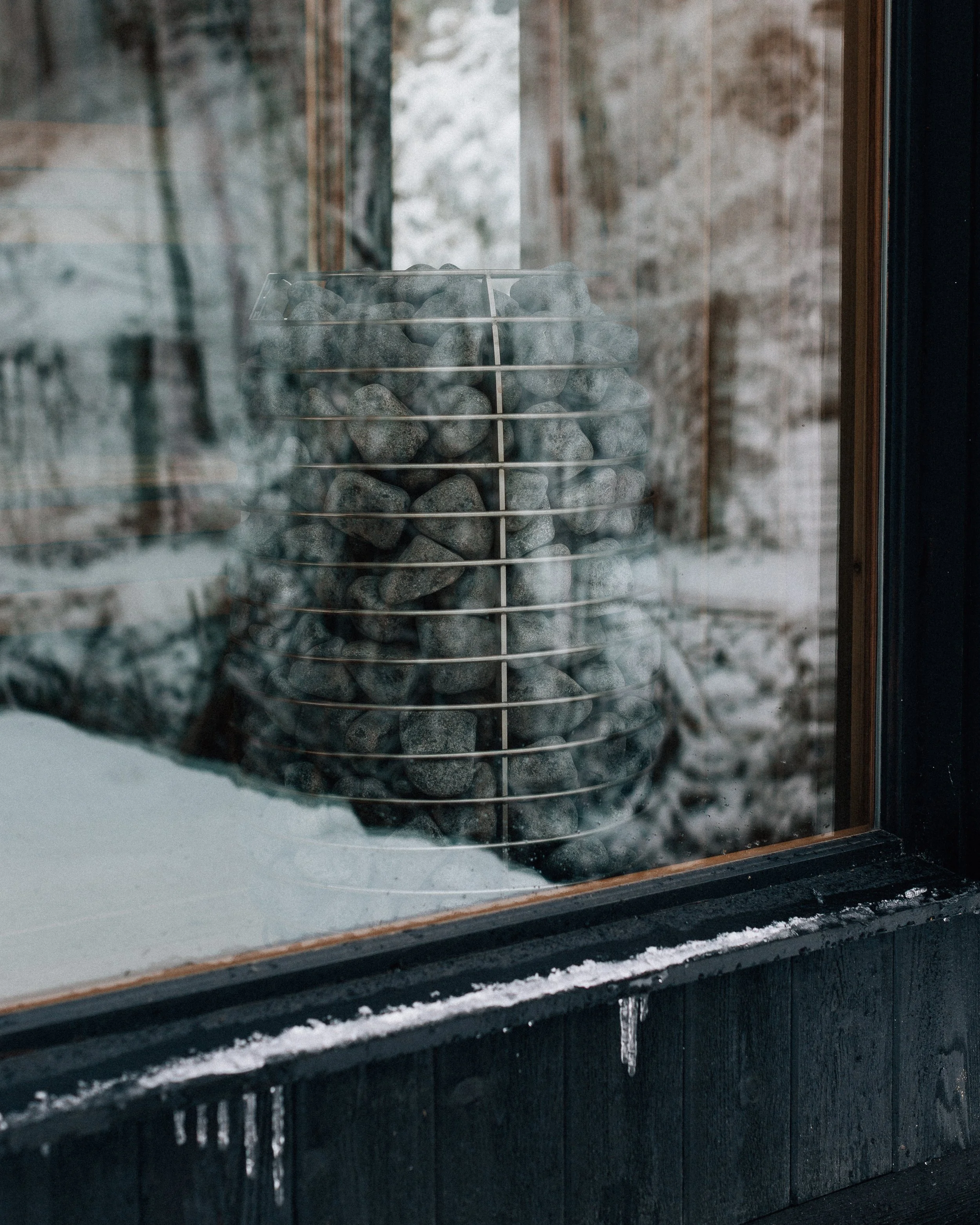 A view through a glass window showing a sauna with a container filled with smooth gray stones, and snow falling outside.