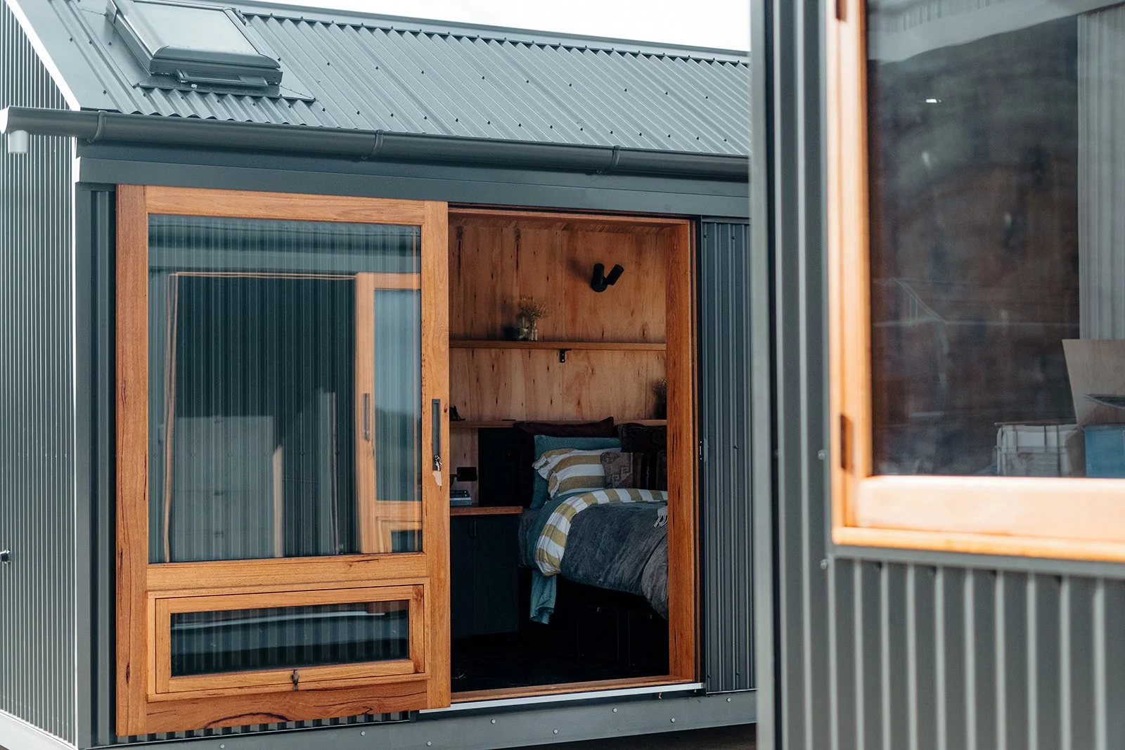 A small modern shelter or tiny house with a snow-covered roof located in a snowy forest with leafless trees.