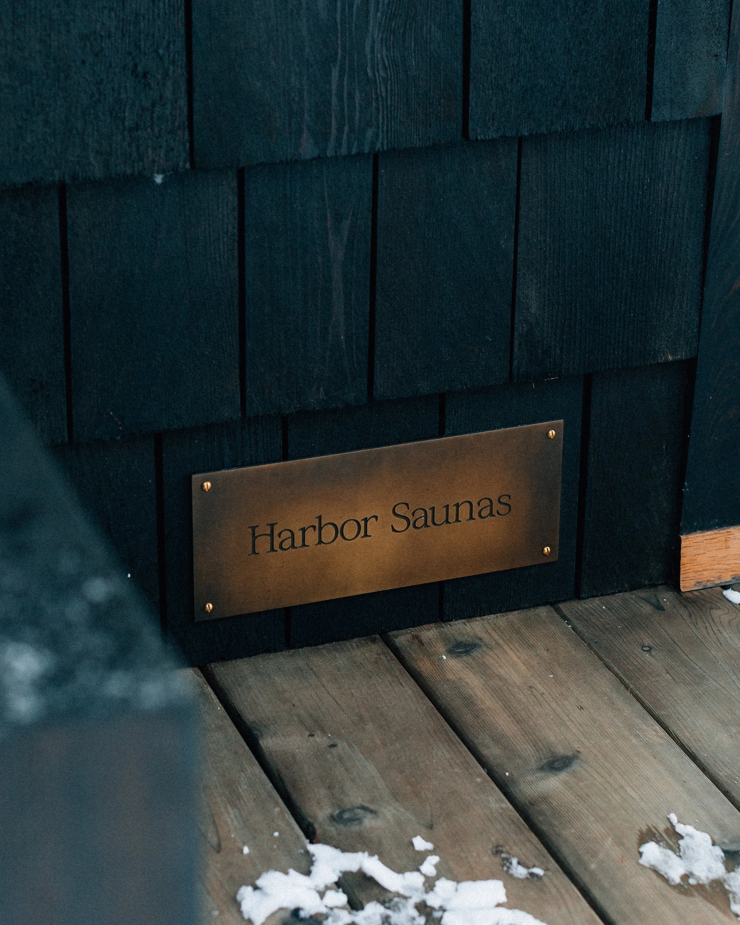 A black wooden wall with a gold-colored sign that reads "Harbor Saunas". The wall is made of horizontal planks, and there are some pieces of snow on the wooden surface below.