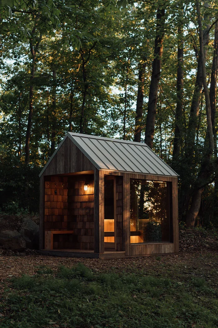 A small wooden outdoor shelter with a sloped metal roof, soft interior lighting, benches, and a wood-fired hot tub, situated in a wooded area during sunset.