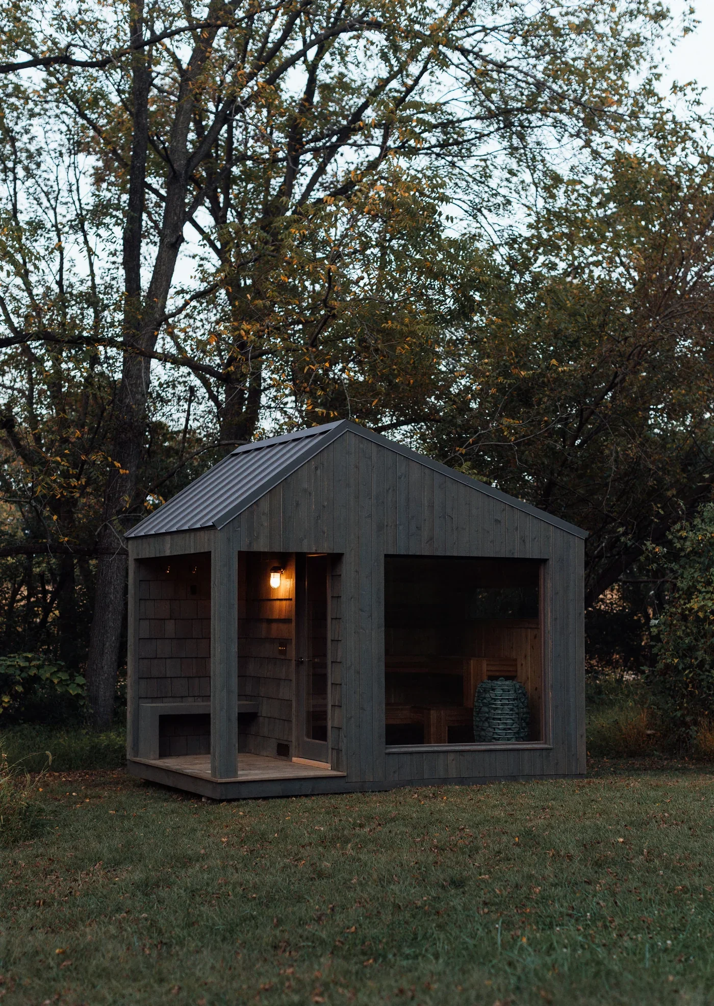 A small wooden building with a sloped metal roof, large windows, and a porch area with a light on inside, situated in a backyard with green grass and surrounded by large trees with autumn-colored leaves.
