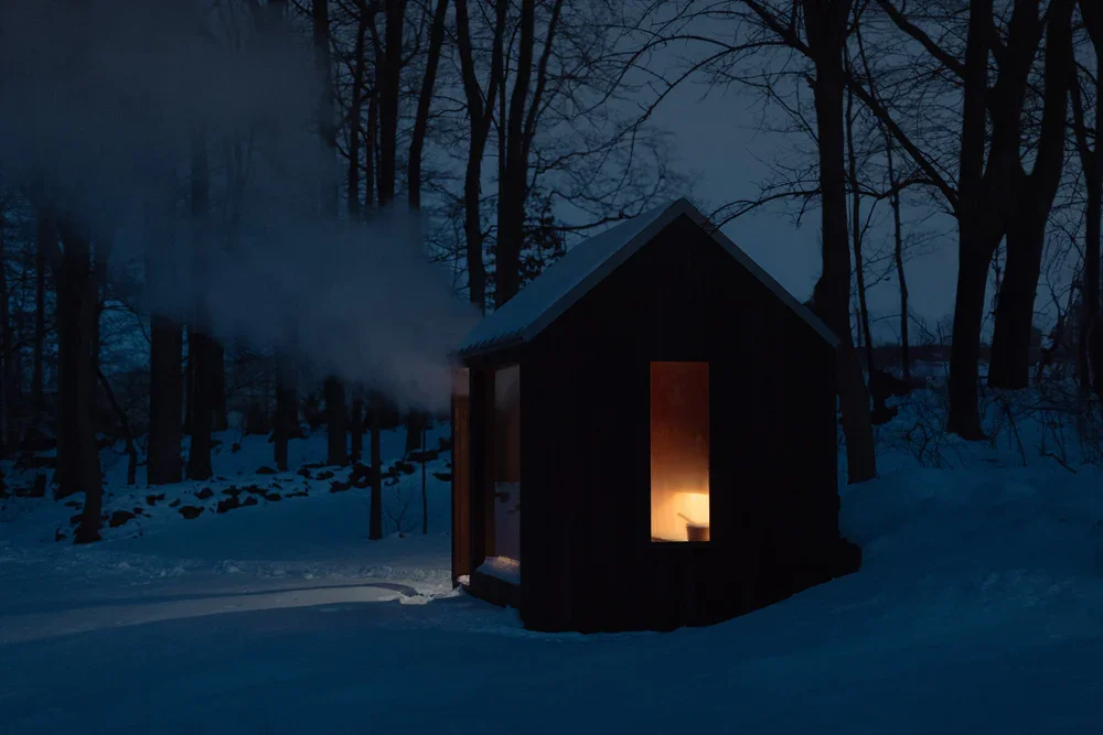 A small, dark cabin in a snowy forest at dusk with warm light glowing inside and smoke coming from the chimney.