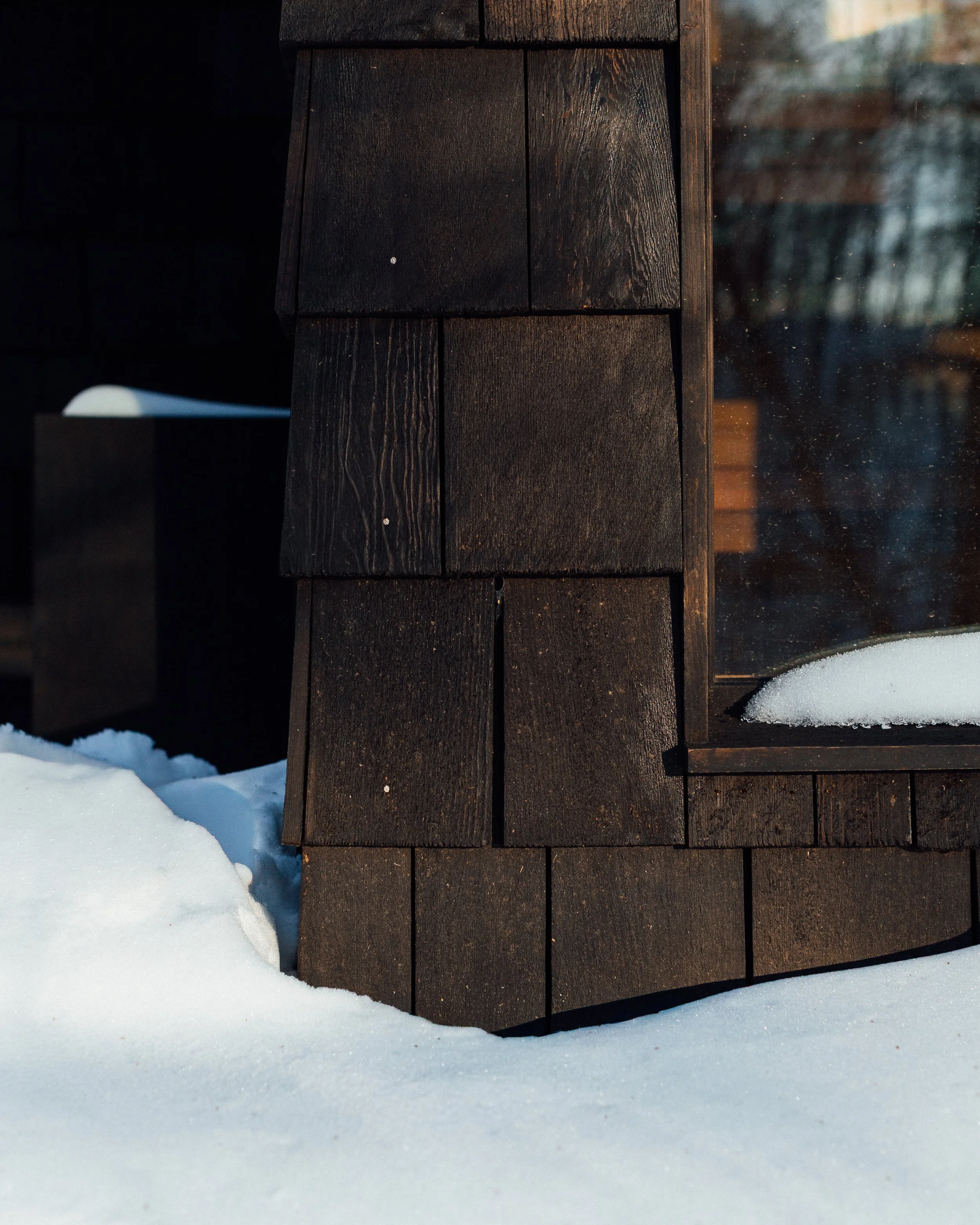 Close-up of a weathered black wooden structure with snow at the base, and a sunlit glass window reflecting trees in the background.