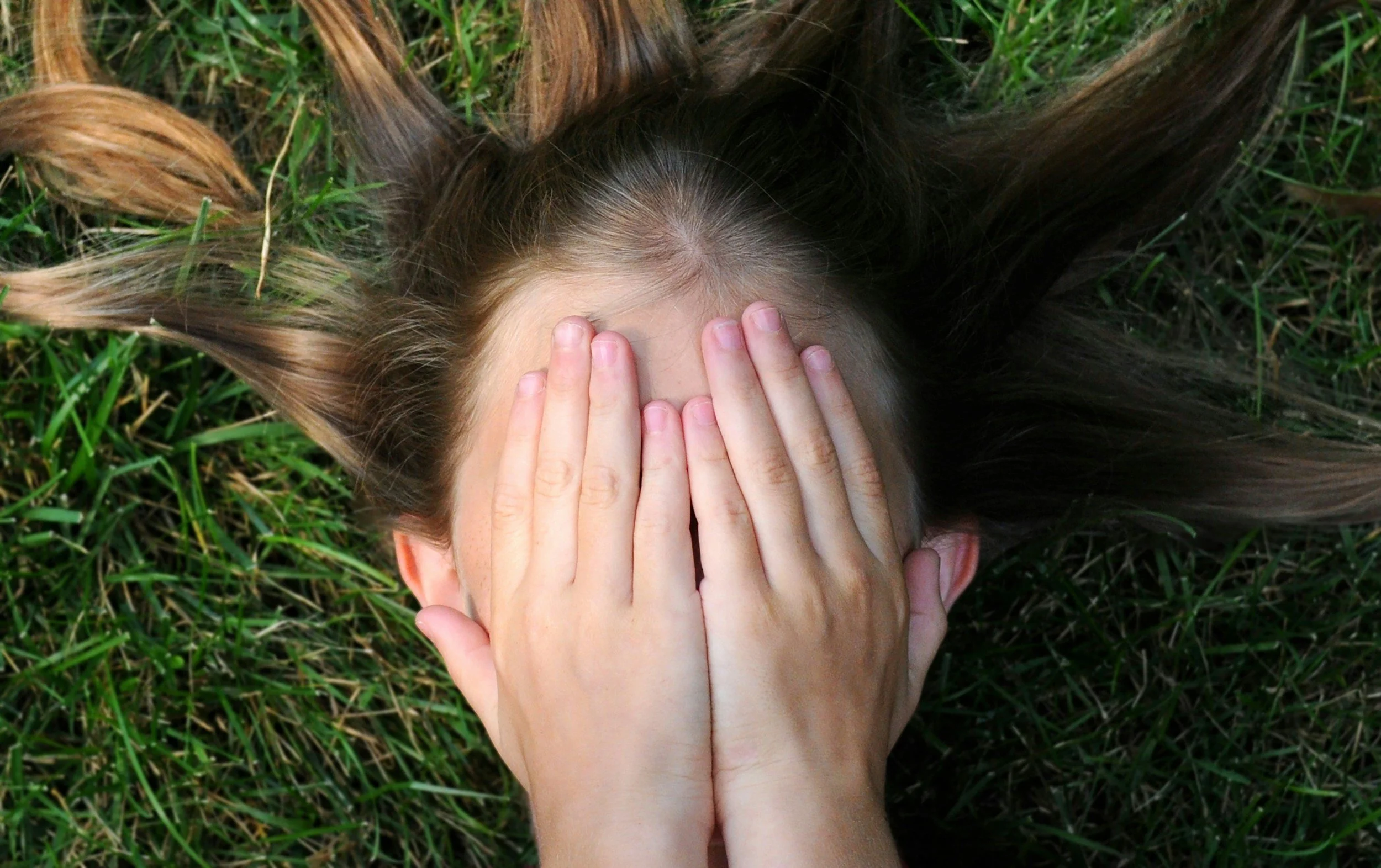 Person lying on green grass with hands covering face, with long brown hair spread around.