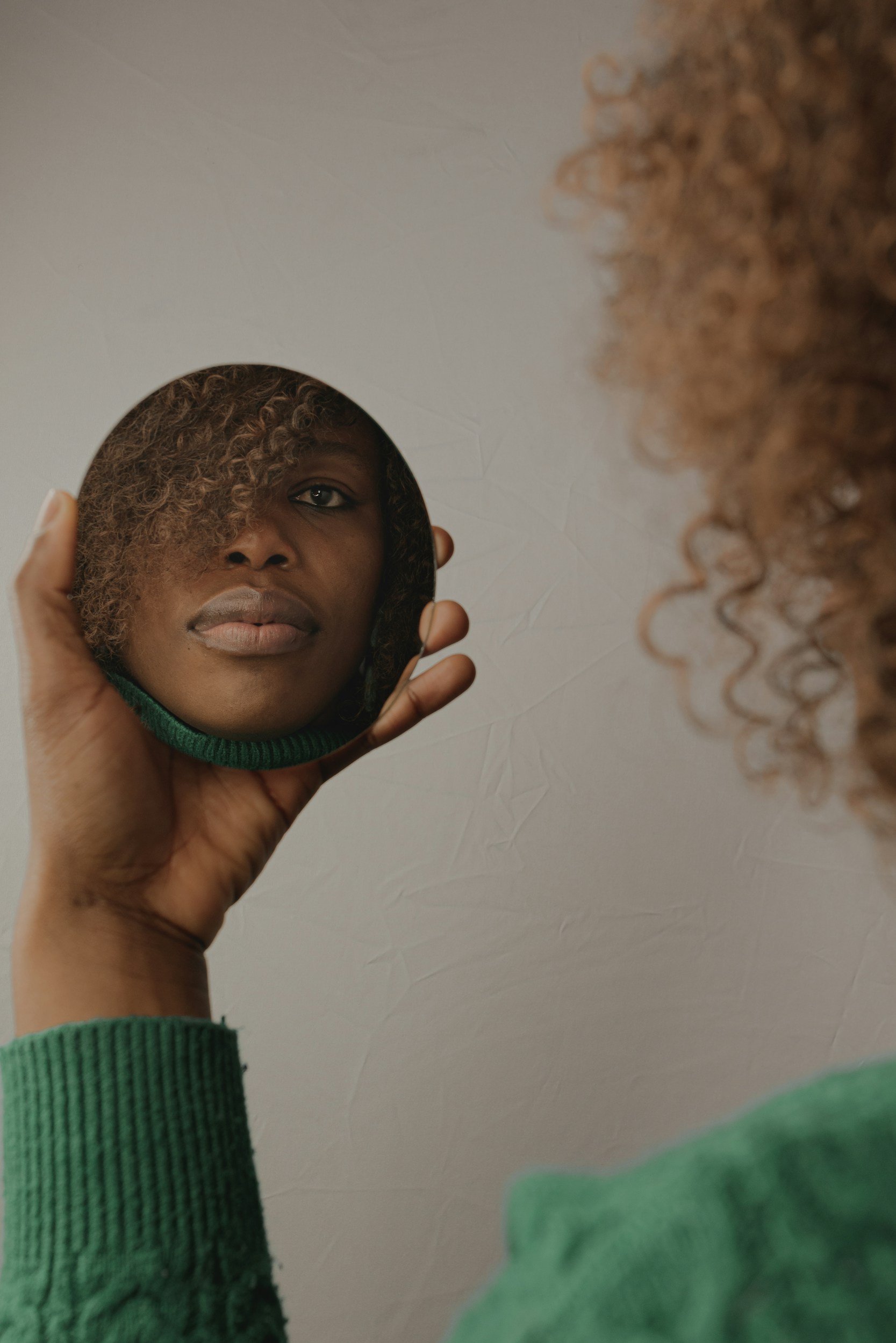 A person holding a small mirror with a reflection of their face, showing curly hair, dark skin, and a green sweater sleeve.