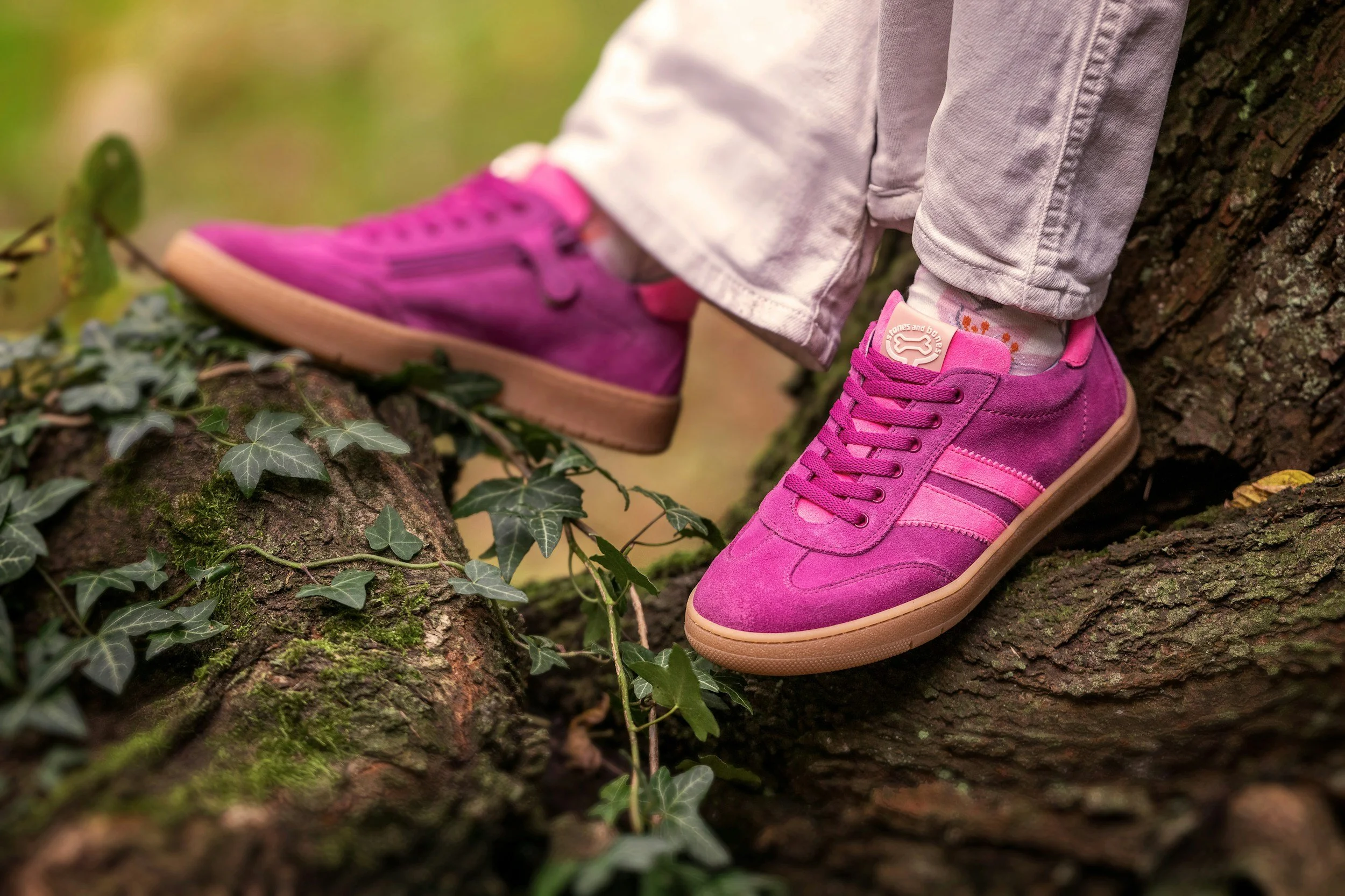 Close-up of a person's pink sneakers and white jeans as they stand on a mossy log in the woods with ivy plants nearby.