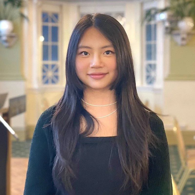 A young man with short black hair and light skin, smiling, standing in front of a brick wall, wearing a plaid shirt and a backpack.