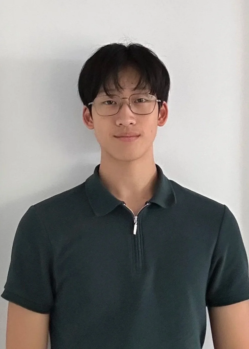 Young man with short black hair and a slight smile standing outdoors, surrounded by green bushes and pink flowers.