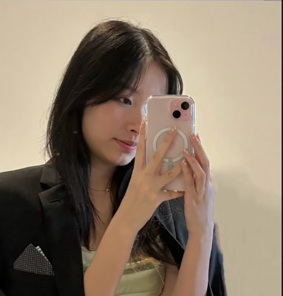 A woman takes a mirror selfie with her smartphone, wearing a black blazer over a light-colored top, with dark hair and a subtle smile.