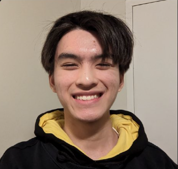 A young man with dark hair smiling at the camera, wearing a black hoodie with a yellow inner lining, standing against a plain indoor background.