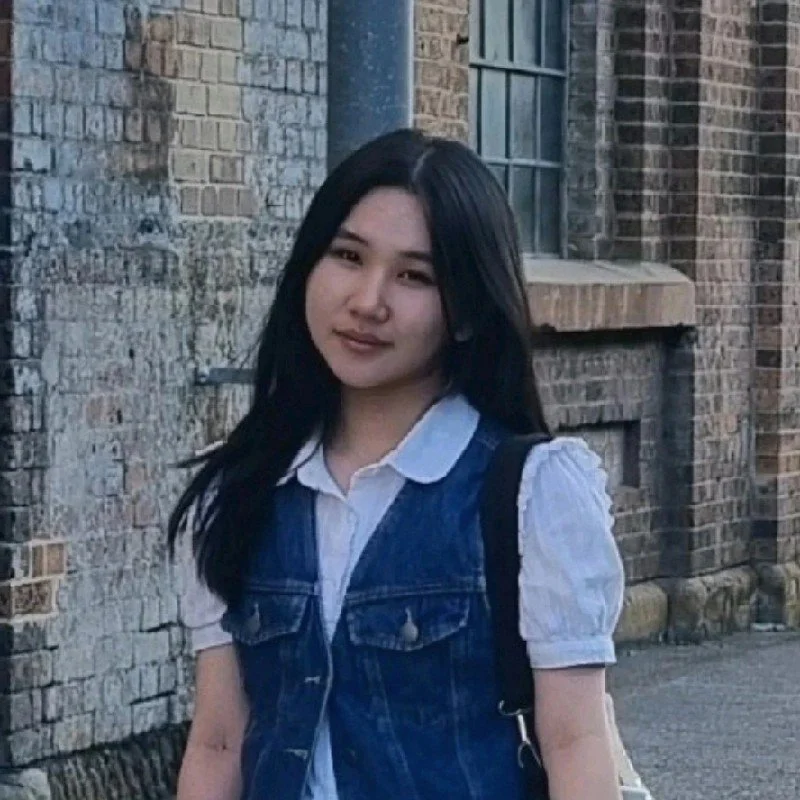 A woman takes a mirror selfie with her smartphone, wearing a black blazer over a light-colored top, with dark hair and a subtle smile.