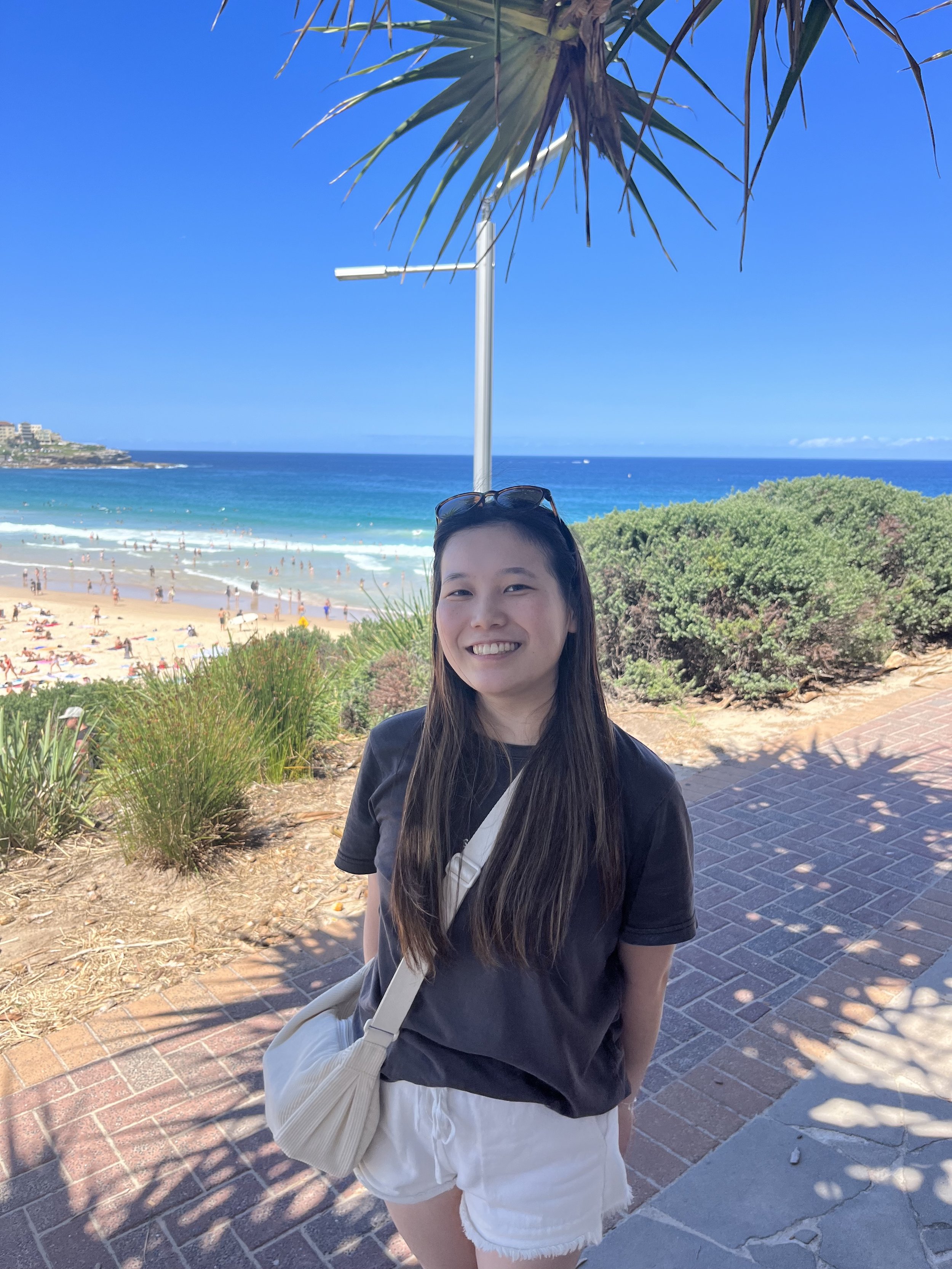 A young woman smiling at the camera on a sunny day at the beach, with the ocean, beachgoers, and blue sky in the background.