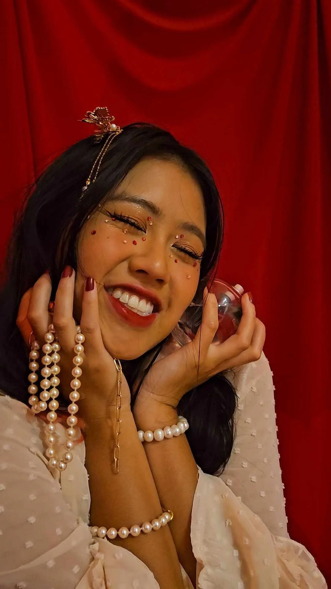 A woman smiling with closed eyes, wearing pearl jewelry, red and gold makeup, and decorative face gems, posing against a red curtain backdrop.
