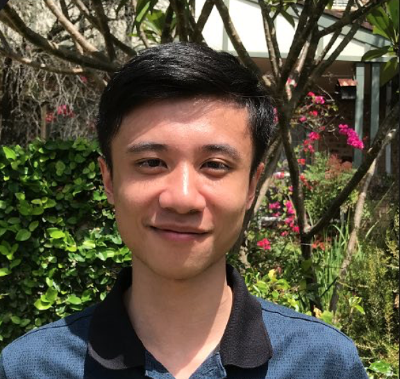 Young man with short black hair and a slight smile standing outdoors, surrounded by green bushes and pink flowers.