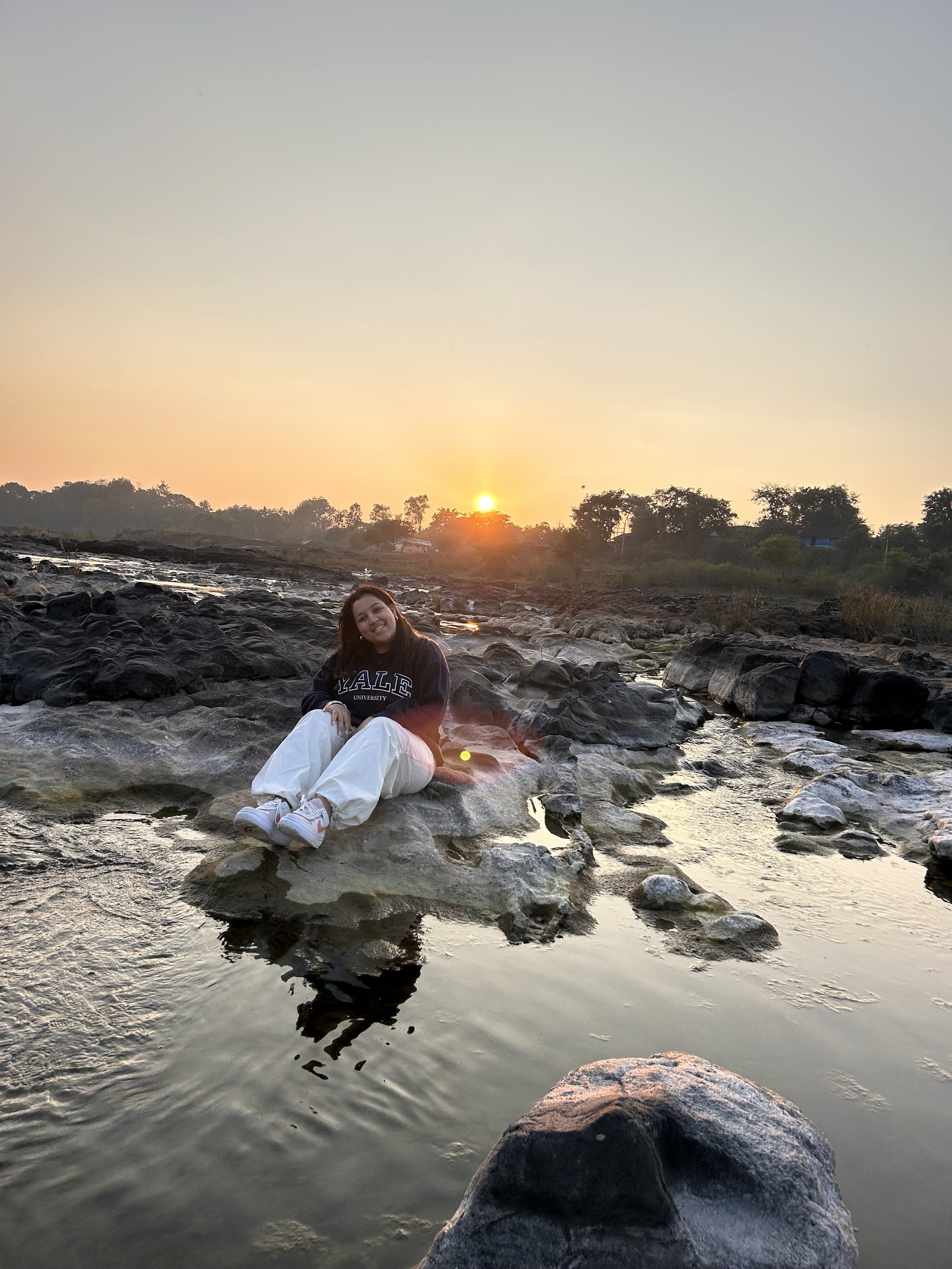 A woman sitting on rocks by a river during sunset, smiling at the camera, wearing a black Yale sweatshirt and white pants.