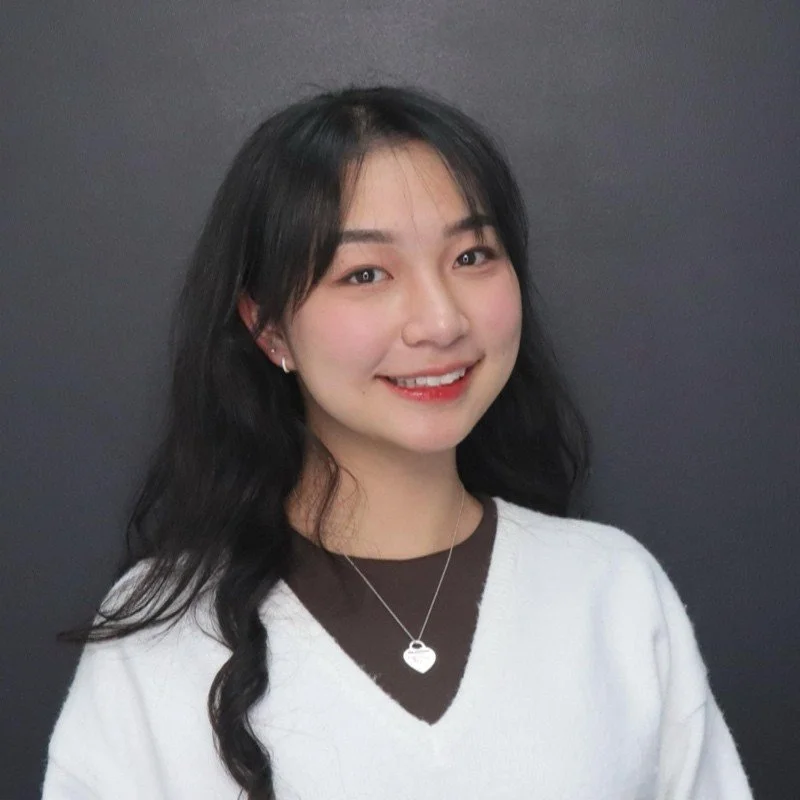A young man with dark hair smiling at the camera, wearing a black hoodie with a yellow inner lining, standing against a plain indoor background.