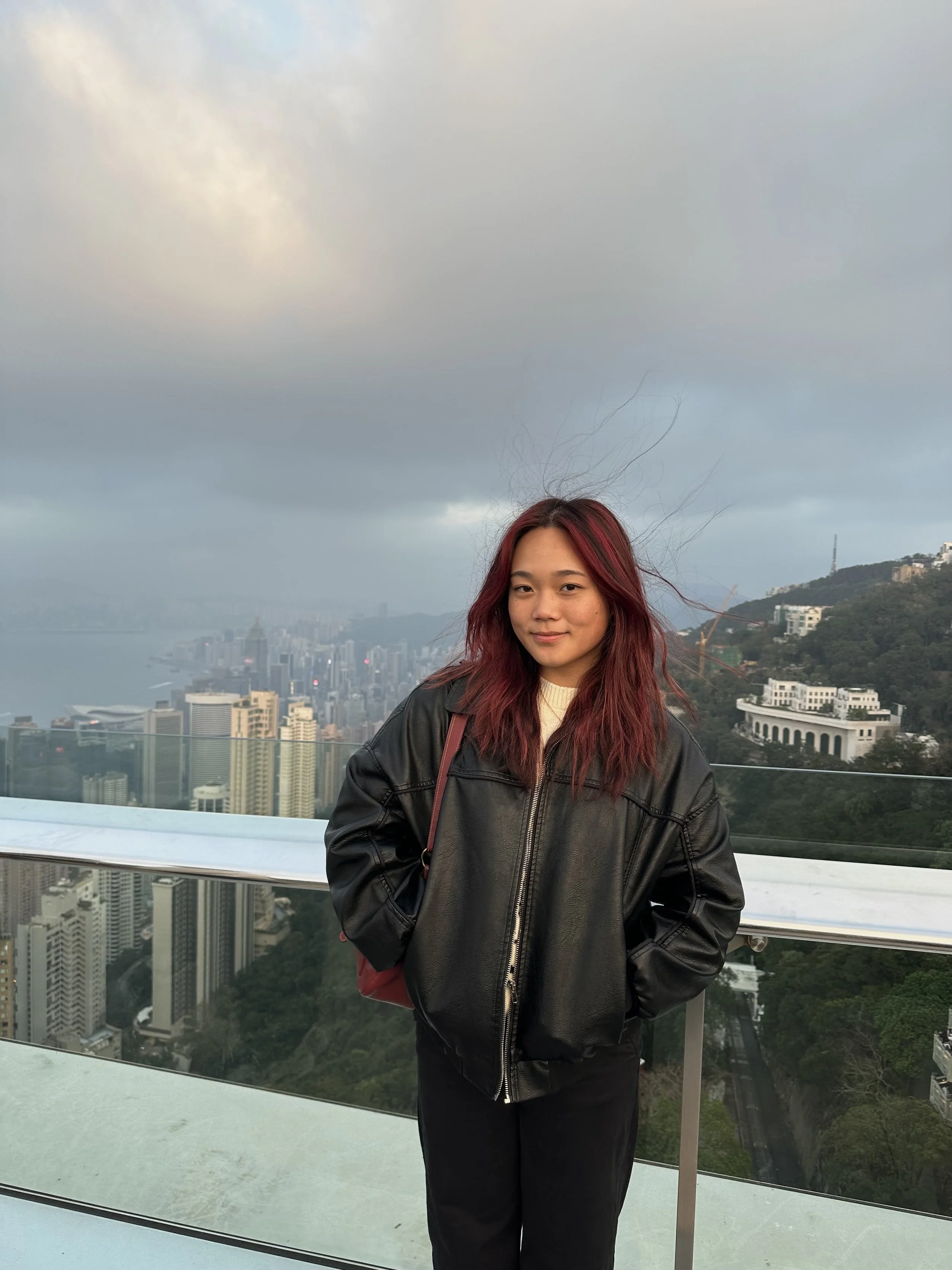 Young woman with red hair wearing a black leather jacket standing on an observation deck overlooking a city skyline and harbor with cloudy sky.