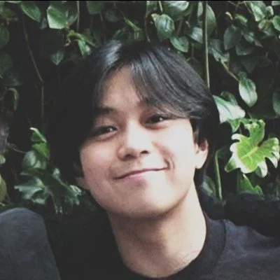 A young person with black hair smiling outdoors surrounded by green leafy plants.