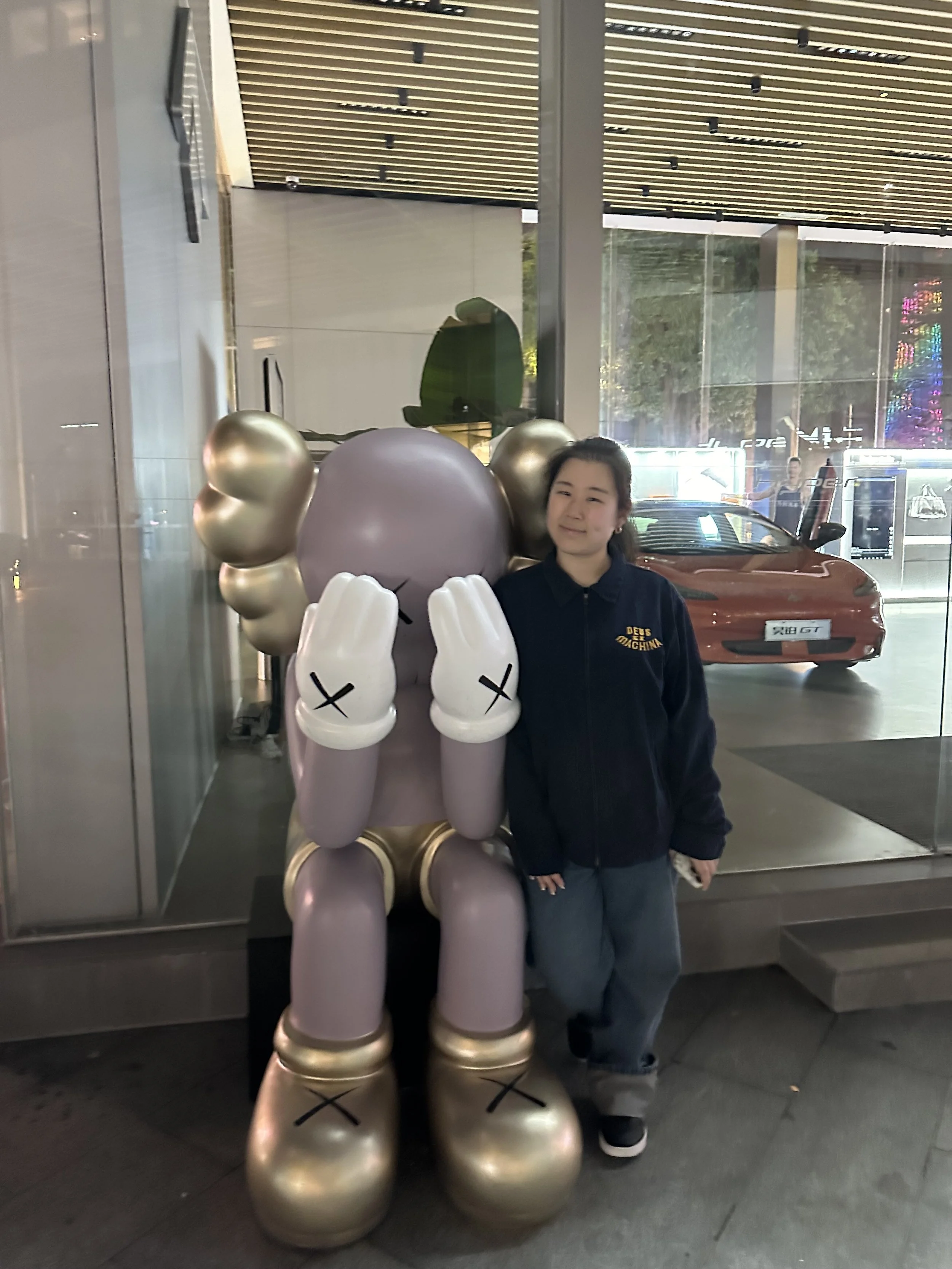 A girl standing next to a large cartoon-like statue of a character with a round head, closed eyes, and hands covering its ears, inside an urban building with a glass window and a view of parked cars outside.