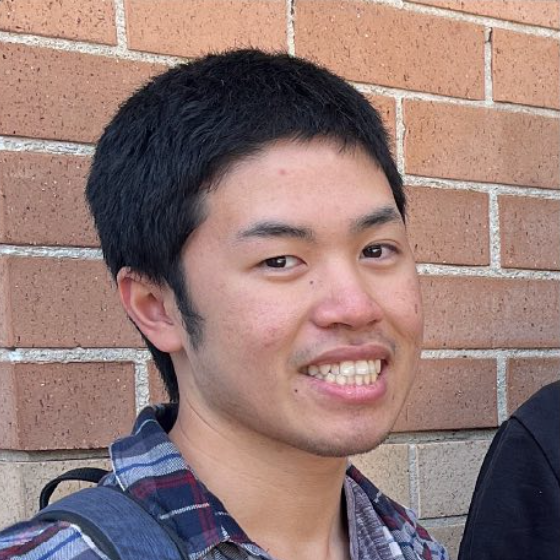 A young man with short black hair and light skin, smiling, standing in front of a brick wall, wearing a plaid shirt and a backpack.