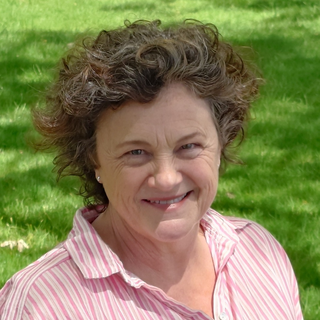 Smiling woman with brown hair wearing a patterned shirt, standing outdoors against a blurred green background.