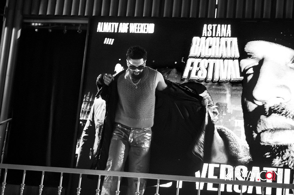 Almaty, Kazakhstan — A black and white photo of a man wearing sunglasses, a sleeveless sweater, and shiny pants, adjusting his jacket in front of a large promotional poster for the Astana Bachata Festival. The poster features a close-up profile of a smiling woman and the