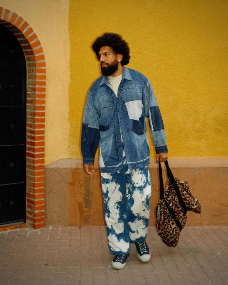 Man wearing a blue patchwork denim jacket and tie-dye pants, holding leopard print bag, standing against yellow wall with brick arch window.