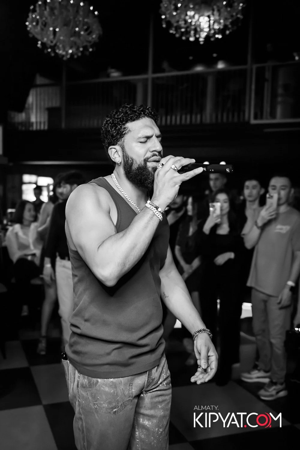 Almaty, Kazakhstan — A man with curly hair, a beard, and jewelry is singing or performing with a microphone at a crowded indoor event. People in the background are watching and recording with their phones.