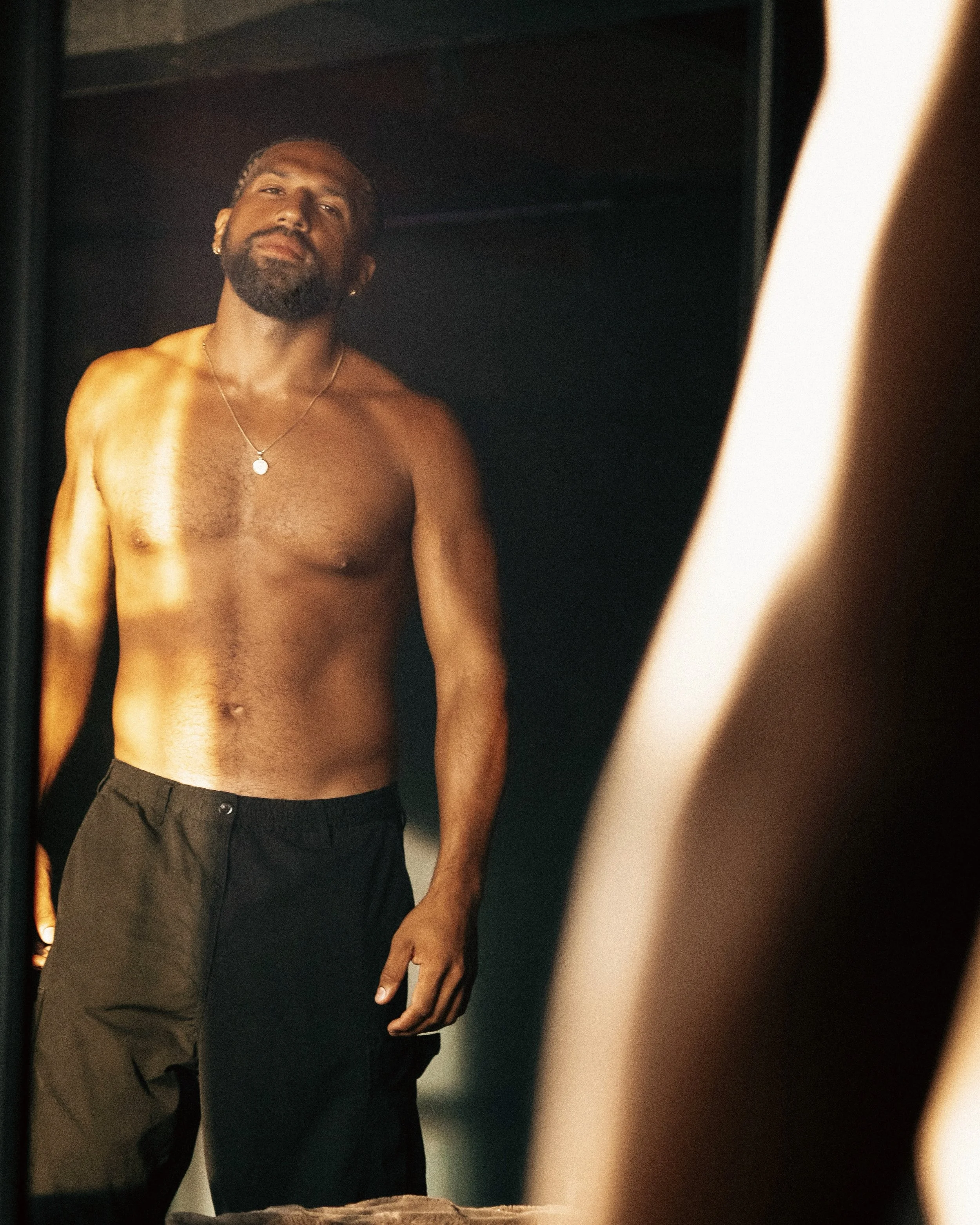 A shirtless man with a beard and necklace standing in front of a mirror, looking at himself.