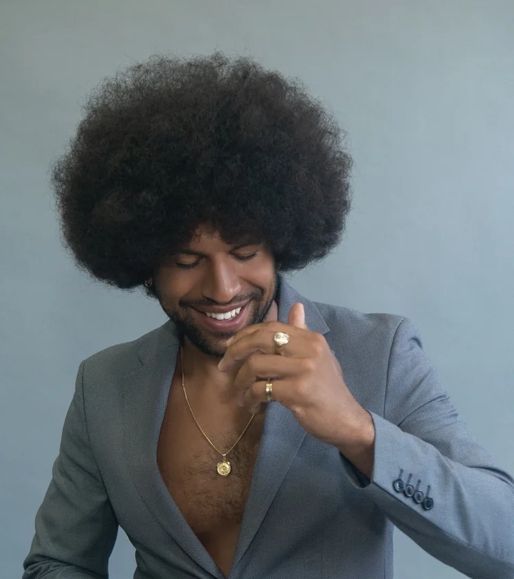 A man with an afro hairstyle wearing a gray blazer, gold jewelry, and rings, smiling and looking down with his hand near his face.
