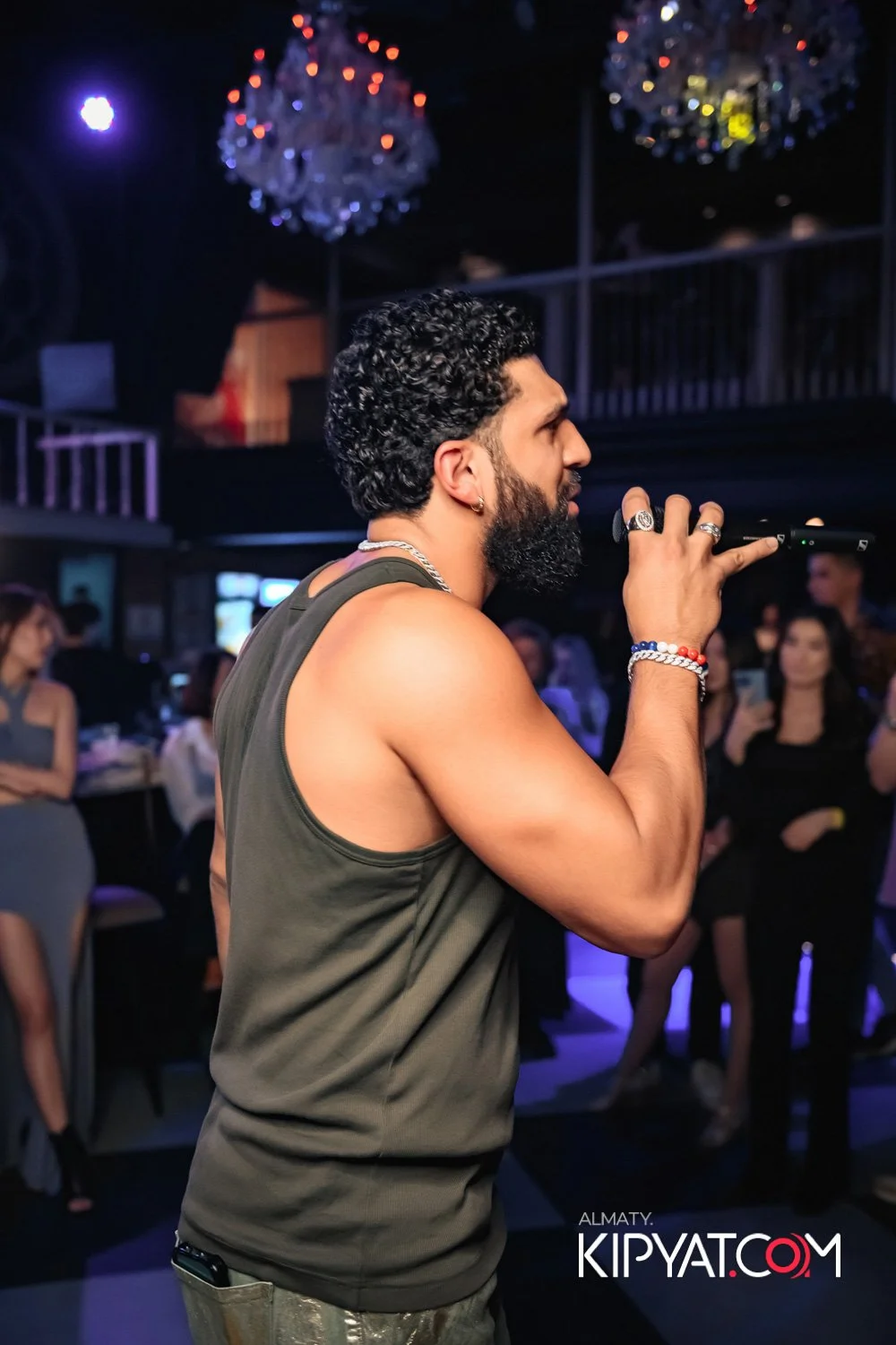 Almaty, Kazakhstan — A man with a curly beard and hair, wearing a sleeveless shirt, holding a microphone and singing or speaking on stage at a nightclub or event, with people in the background watching and taking photos.