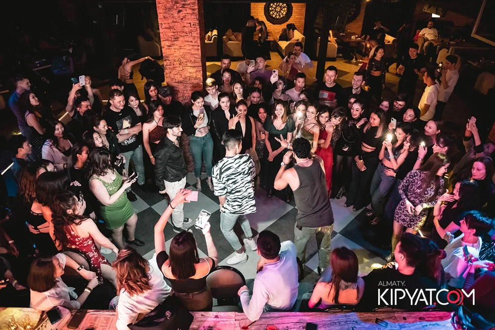 Almaty, Kazakhstan — A large group of people dancing and socializing in a club or bar with dim lighting and brick walls. Some people are taking photos or videos with their phones, and two individuals are performing or leading a dance in the center of the crowd.
