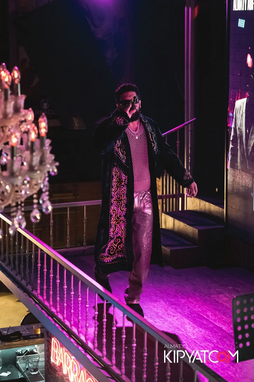 Almaty, Kazakhstan — Man on stage with dark sunglasses, wearing a long embroidered coat, a pink mesh top, and shiny pants, illuminated by purple and pink stage lighting, with a chandelier and stairs in the background.
