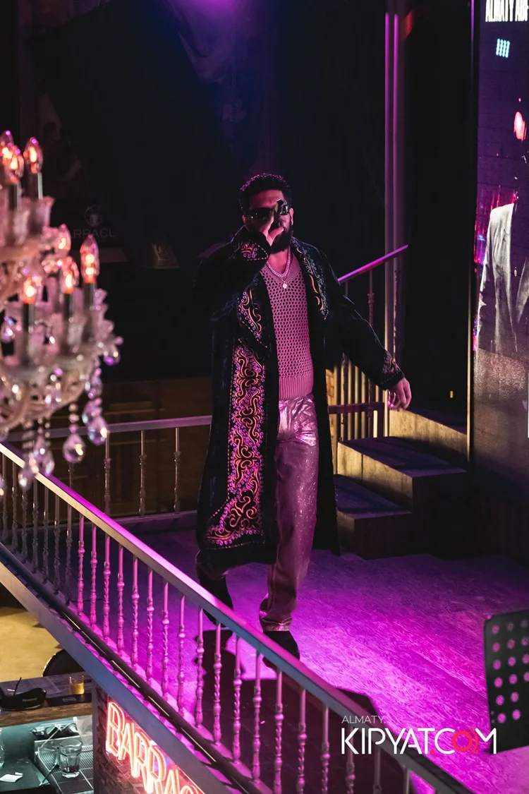 Almaty, Kazakhstan — Man on stage with dark sunglasses, wearing a long embroidered coat, a pink mesh top, and shiny pants, illuminated by purple and pink stage lighting, with a chandelier and stairs in the background.