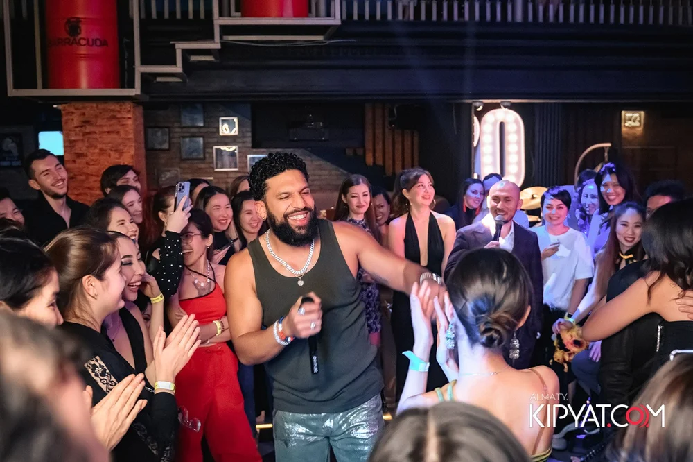 Almaty, Kazakhstan — A man with a beard and curly hair, wearing a black sleeveless shirt and silver jewelry, is laughing and dancing among a crowd of smiling women and men at an indoor party or event, with a large illuminated letter and brick walls in the background.