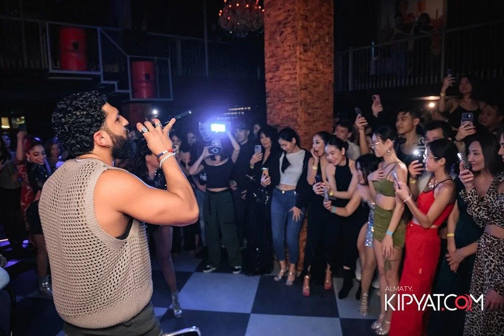Almaty, Kazakhstan — A man with curly hair, beard, and sleeveless beige sweater singing into a microphone on stage at a nightclub, while a crowd of young women in stylish outfits watch and record with smartphones.