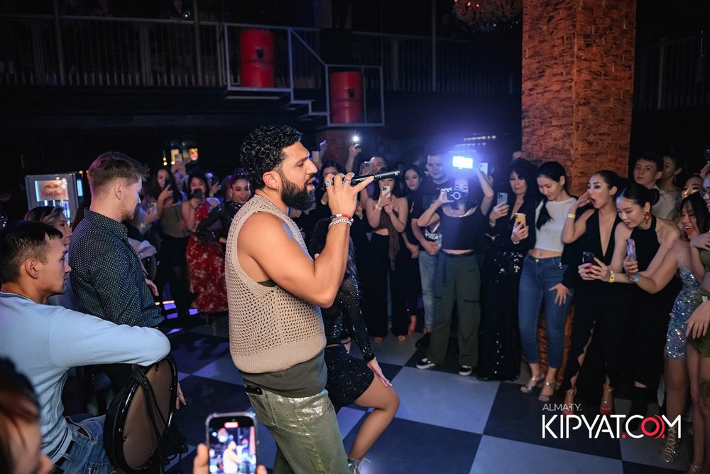 Almaty, Kazakhstan — Man with curly hair and beard singing into microphone while surrounded by a crowd at a nightclub or party