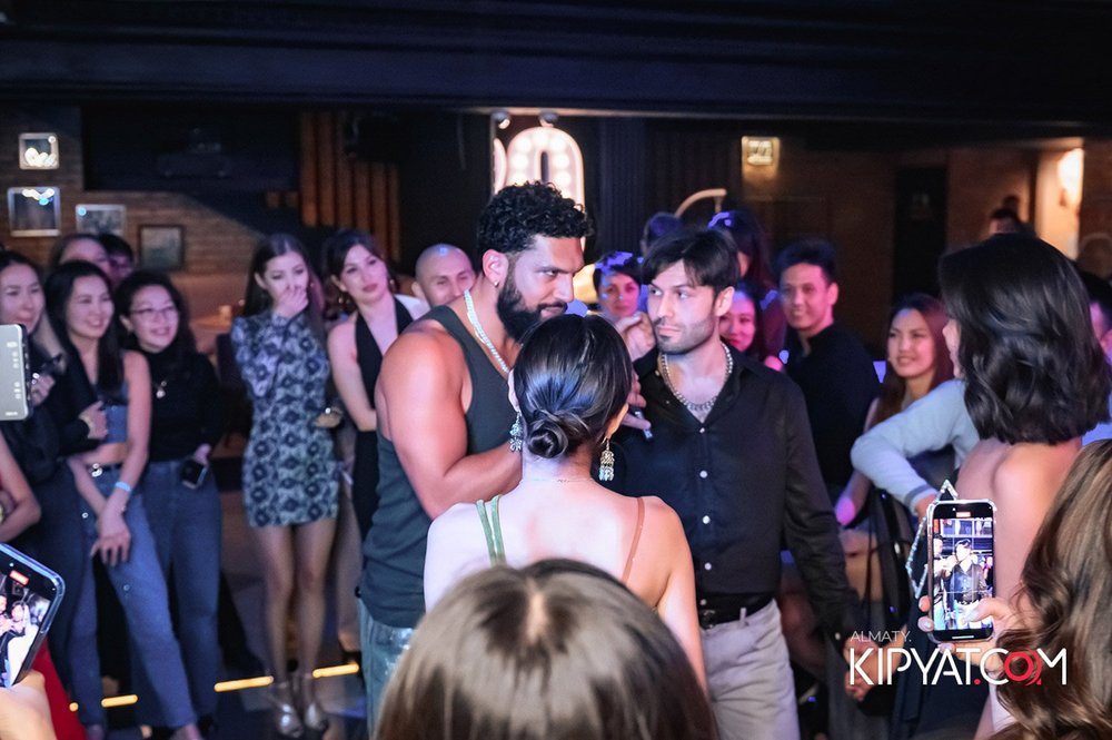 Almaty, Kazakhstan — A group of people at a lively indoor event. In the center, a muscular man with a beard, wearing a black tank top, appears to be speaking or performing. Two other men and a woman are also near him, engaged in conversation. The background features an a