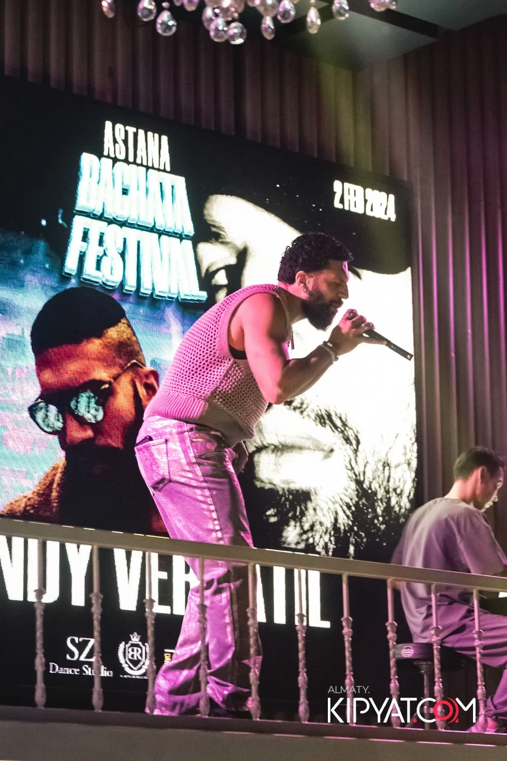 Almaty, Kazakhstan — Performer singing on stage at Astana Bachata Festival 2023 with a large screen behind showing a black and white image of a woman and a man. The stage has a curtain backdrop and stage lights. The performer has dark, curly hair, a beard, and is wearing