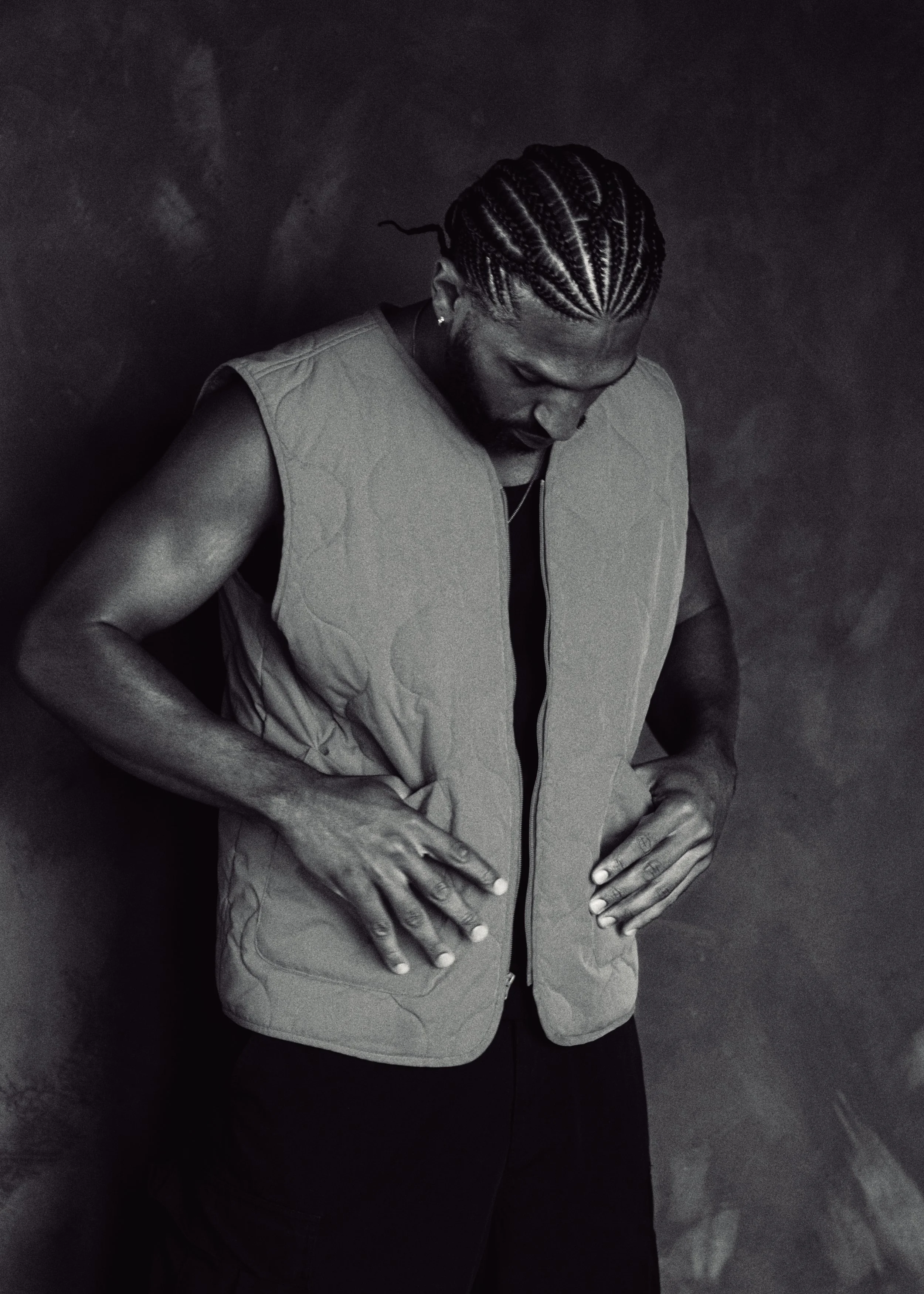 A man with neatly braided hair looking down, wearing a sleeveless quilted vest, standing against a dark background.