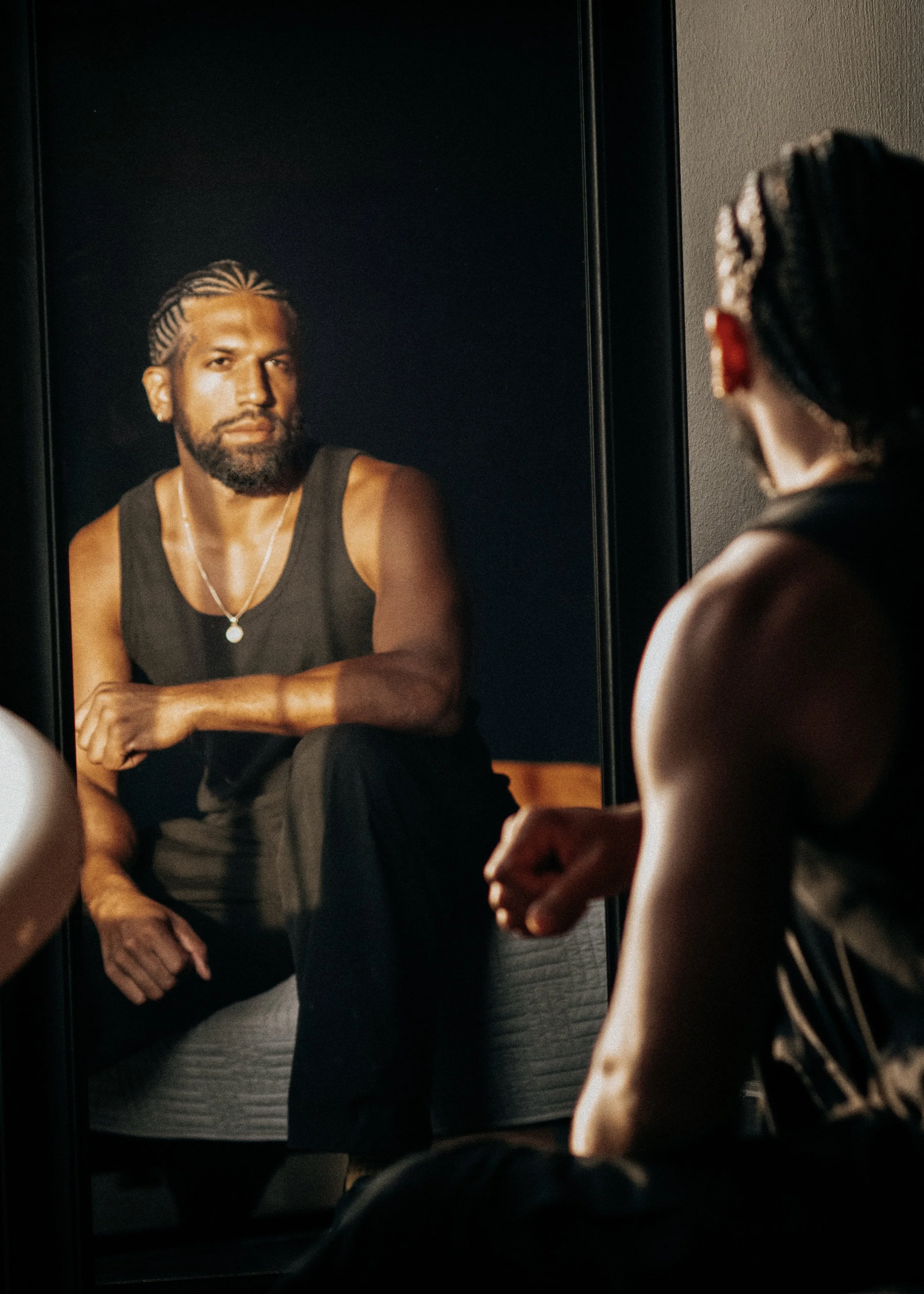 Person with braided hair and earrings looking at a mirror showing a photo of a bearded man with braided hair wearing a black tank top and a silver necklace.