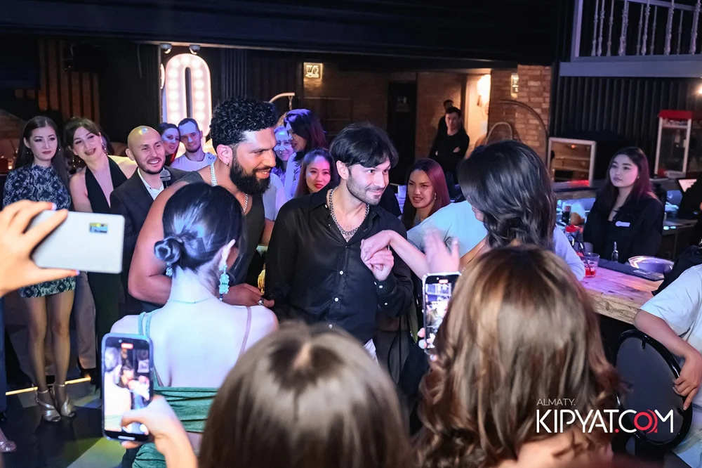 Almaty, Kazakhstan — Group of people at a social event, some are taking pictures, others are smiling and chatting, with a DJ booth in the background.