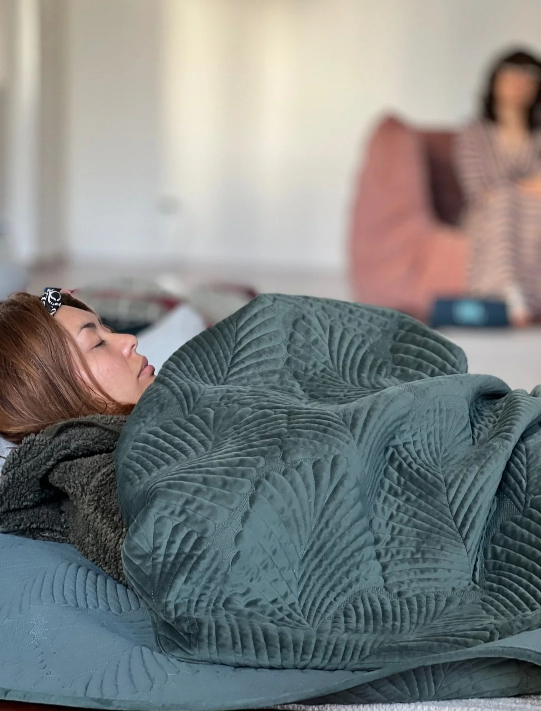 A woman sleeping on a bed, covered with a green quilt, with another woman in the background sitting on the bed.