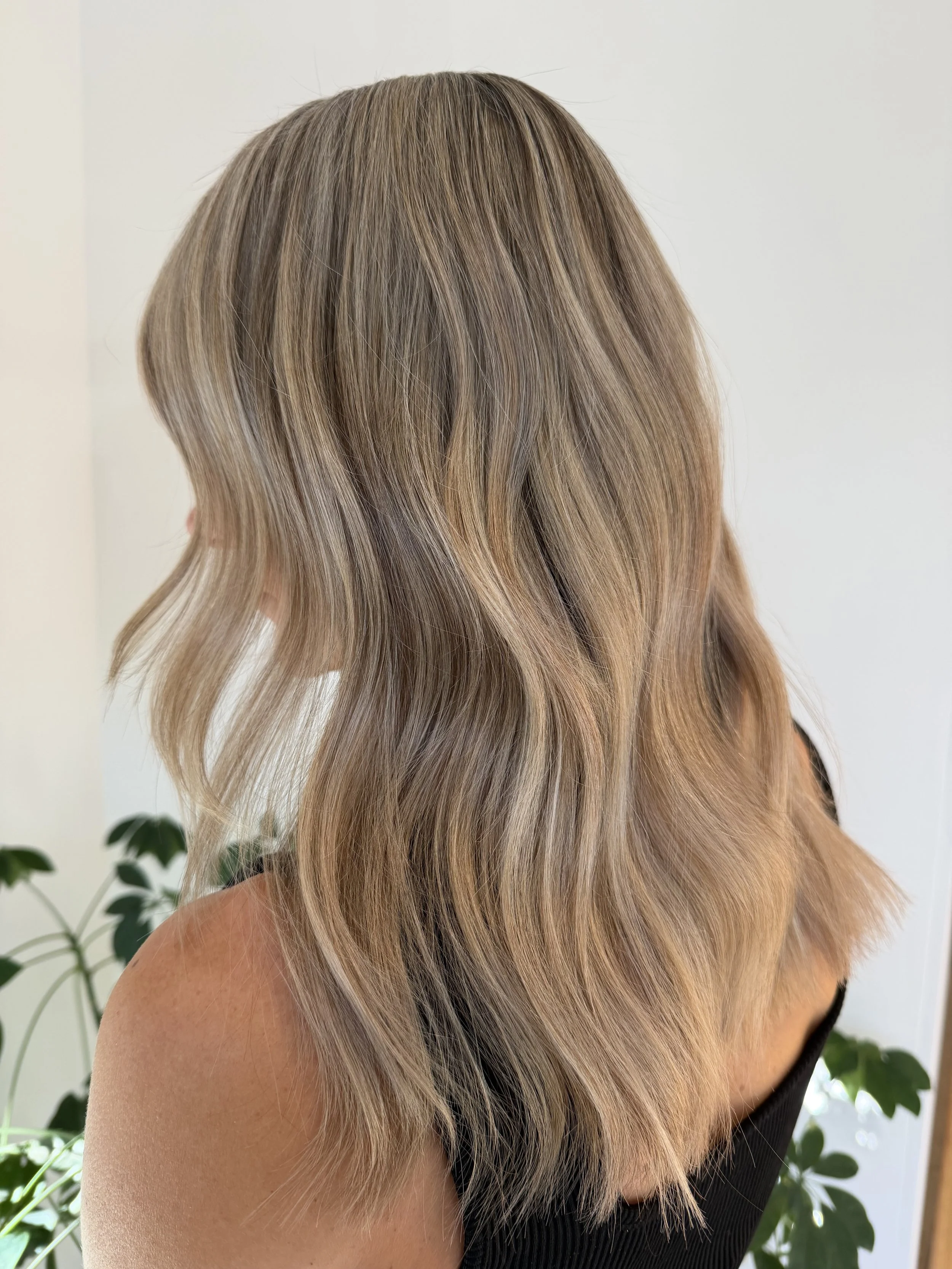 Woman with shoulder-length wavy blonde hair, wearing a black top, standing indoors near a plant and a white wall.
