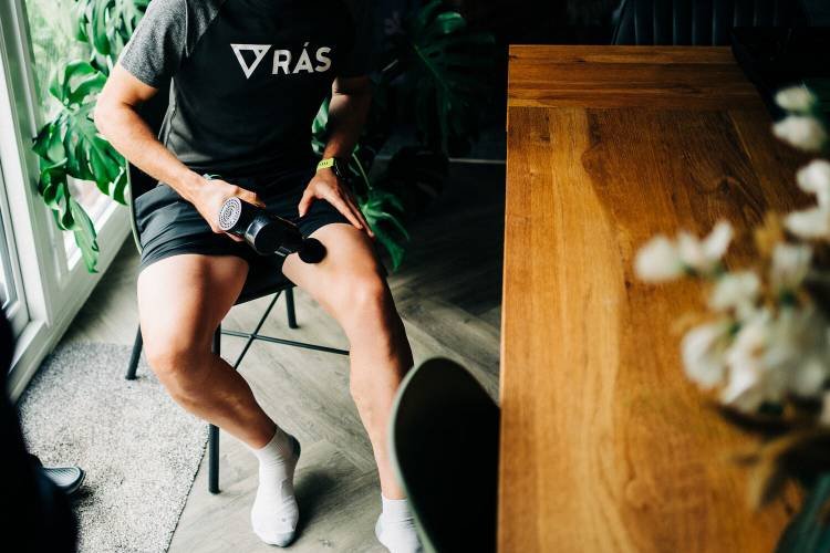 RÁS - Young male sat down by a table using a massage gun on leg muscle