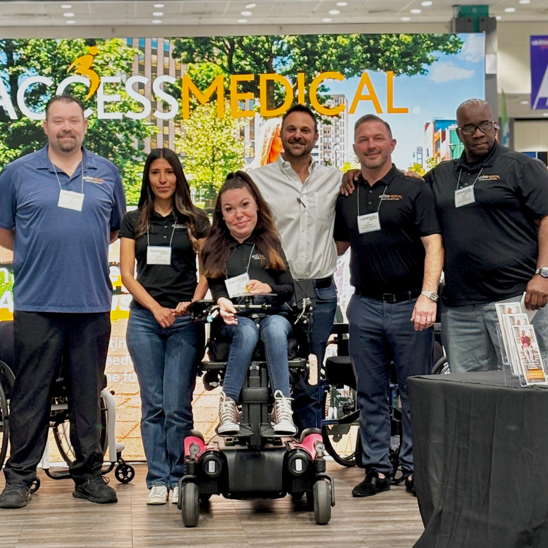 Group of six people standing on stage, with one woman in a wheelchair in front, in front of a backdrop with the logo 'ACCESS MEDICAL' and cityscape background.