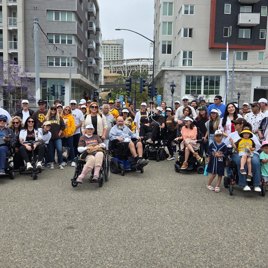 A large group of diverse people, including children, adults, and seniors, gathered outdoors in an urban area with high-rise buildings background, some in wheelchairs and others standing, posing for a group photo.